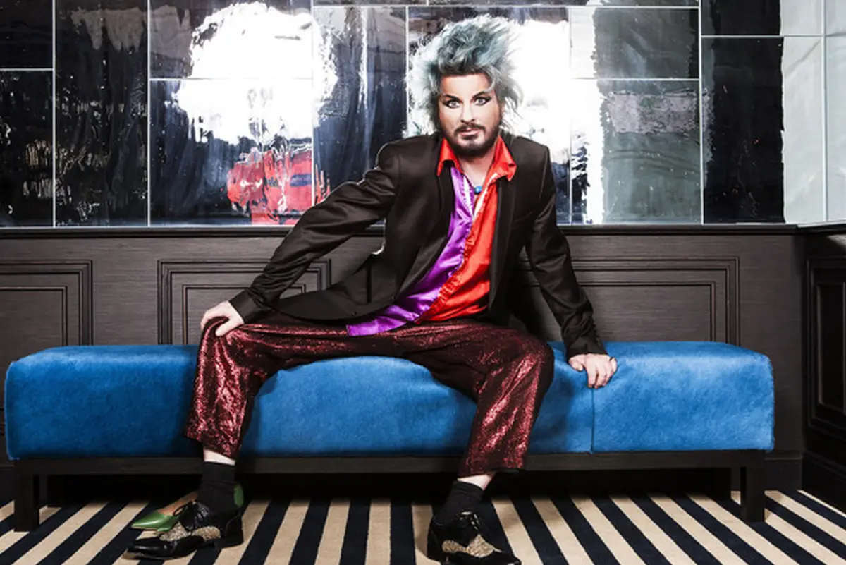 ay Ray sitzt in farbenfroher Kleidung auf einem blauen Sofa.